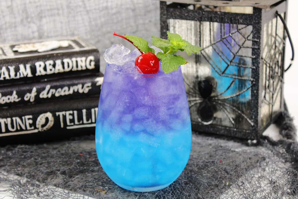 glass with disney themed cocktail that is blue and purple and topped with a maraschino cherry
